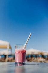 Glass with strawberry milkshake and a paper straw on a table,  at the beach in bright sun light.