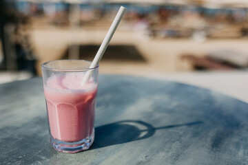 Glass with strawberry milkshake and a paper straw on a table, outdoors in sun light.