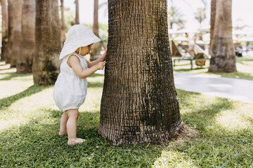 Little child standing barefoot on green grass next to a tree, playing with tree bark.