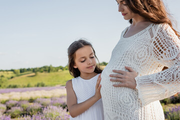 smiling girl hugging tummy of pregnant mother in blurred field.