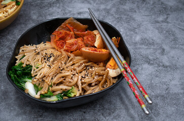 Korean vegetarian noodle bowl with asian mushrooms, bok choy stir fry, kimchi and pickled eggs. Black bowl,  steel chopsticks on gray table. Copy space.