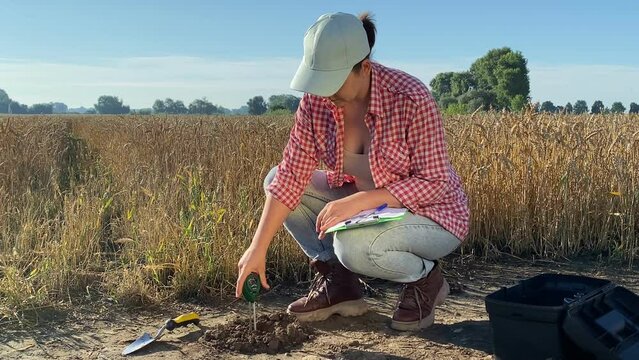 Female agriculturist measuring soil features by using soil tester, writing data in information sheet at sunrise outdoors. Professional woman farmer applying measurement instrument at agricultural