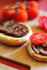 The hamburger is being prepared and served with sliced tomatoes, onions, pickles. on a wooden cutting board in the kitchen