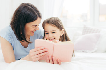 Fototapeta premium Indoor shot of pleasant looking female child with curious expression, reads interesting book together with her affectionate mother, views colourful pictures, lie on bed in spacious bedroom. Childhood