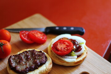 The hamburger is being prepared and served with sliced tomatoes, onions, pickles. on a wooden cutting board in the kitchen