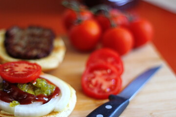 The hamburger is being prepared and served with sliced tomatoes, onions, pickles. on a wooden cutting board in the kitchen