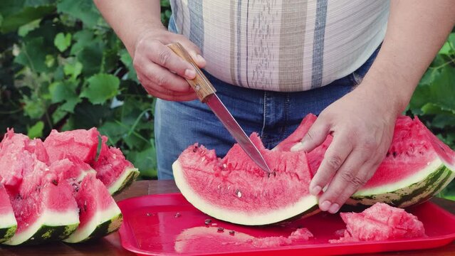 A man carefully removes the bones from a red slice of watermelon with a knife. Pieces of delicious ripe watermelon lie on the table ready to eat. Camping, picnic in the garden or cottage. UHD 4K.