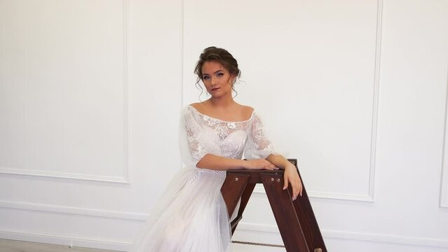 A charming girl in a wedding dress poses leaning on a bar. Charming young brunette bride on a light isolated background in a white dress. Space for copy. UHD 4K.