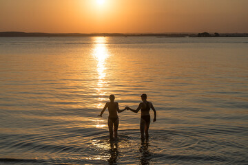 Fototapeta premium Couple at sunset in the water. A happy couple of lovers walk along the beach on a beautiful summer evening. Romance, relationship, love concept.