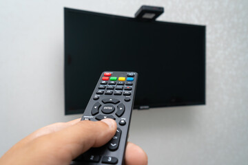 Close-up of a remote control in the hand of a man directing at a black LCD TV hanging on a white wall