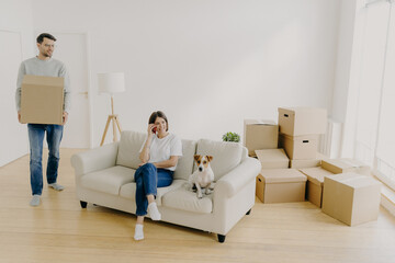 Loving spouses pose in rented apartment, woman relaxes on couch with pet, speaks via modern...
