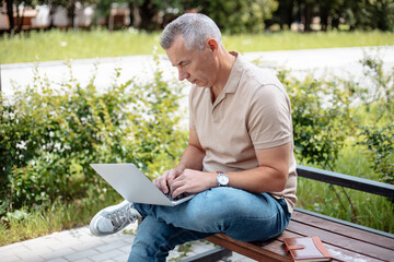 Portrait of concentrated elderly man sitting on bench with crossed leg in park in summer, working...