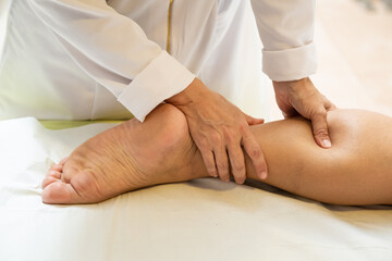 Uma terapeuta vestida de branco, fazendo massagem na perna de paciente deitado em maca. - obrazy, fototapety, plakaty