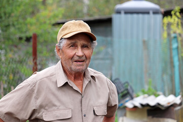 Portrait of elderly man in baseball cap and short sleeve shirt standing in rural yard. Life in...