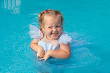 Fototapeta na wymiar Charming girl 3 years old in a blue dress with blond hair in the pool smiles. Summer holiday concept. Place for text