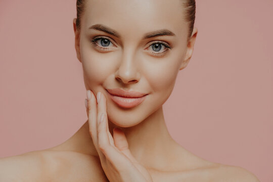 Cropped shot of attractive calm woman touches face gently, has direct gaze at camera, cares about skin, has well groomed skin, enjoys facial skin care, stands naked against pink studio background