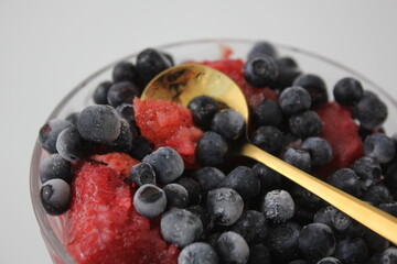  Frozen blueberries and strawberries in a transparent bowl with a spoon on a white background.