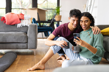 Happy biracial lgbt male couple moving house, sitting on floor in living room, using smartphone