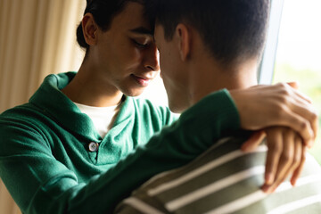 Happy biracial lgbt male couple embracing and smiling next to window