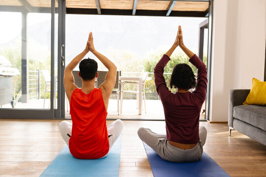 Happy biracial lgbt male couple doing yoga in living room, meditating