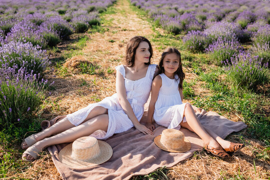 woman and girl in white dresses sitting on blanket in meadow near straw hats.