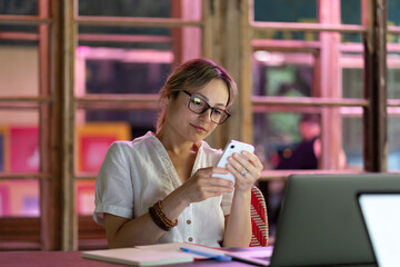 Female employee using mobile device, young woman freelancer distracted by smartphone, sitting at desk with laptop. Freelance procrastination, smartphone distraction at work, workplace distractions