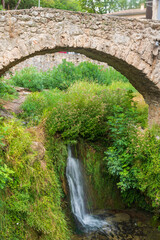 Fototapeta na wymiar Petite chute d'eau dans une nature verdoyante surmontée par un pont médiéval en pierres à Saint-Guilhem le Désert, en Occitanie