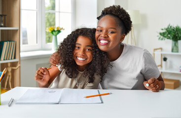 Family having fun while doing homework. Happy parent helping child with school test. Cheerful Afro American daughter and mother sitting at desk with notebook and pencils, looking at camera and smiling