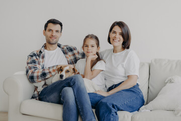 Photo of happy friendly family of mum, dad and daughter pose on comfortable sofa with pet, have good relationship, dressed casually, smile positively. Family time and domestic atmosphere concept