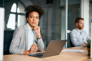 Black businesswoman working on laptop at corporate office and looking at camera.