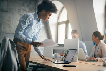 Black businesswoman analyses paperwork while working on computer in office.