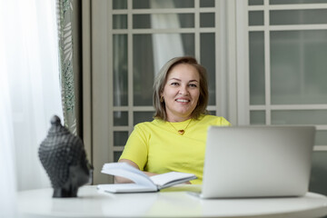 attractive blonde middle-aged woman entrepreneur in a yellow blouse working on a laptop and with documents at home at the table