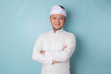 Portrait of a confident smiling Balinese man standing with arms folded and looking at the camera isolated over blue background, wearing a blue shirt