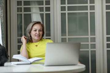 attractive blonde middle-aged woman entrepreneur in a yellow blouse working on a laptop and with documents at home at the table