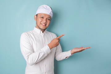 Excited Balinese man wearing udeng or traditional headband and white shirt pointing at the copy space beside him, isolated by blue background
