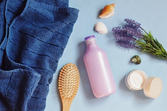 Blue towel, wooden comb, purple shampoo bottle, face cream, lavender flowers and face cream on a blue background. Hair care cosmetic products flat lay photography