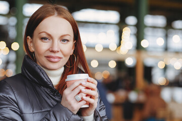 Outside shot of satisfied attractive female model with healthy skin, brown hair, holds takeaway coffee, enjoys good drink, dressed in jacket, waits for someone, has coffee break, looks at camera