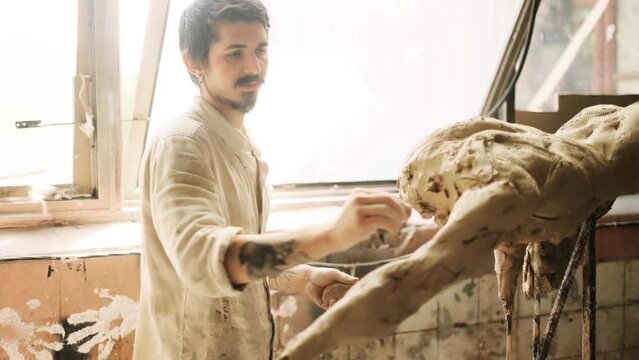 Young bearded man sculpting statue of beautiful woman body in natural light from window.