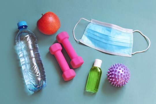 Return to exercise after COVID. Water bottle, apple, pink dumbbells, hand sanitizer and face mask on a green background. Fitness flat lay image