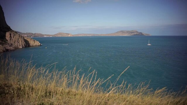 view of the sea from the high bank, where already dry grass sways on a summer evening. a small boat is sailing on the sea and a view of the high mountains on the left.