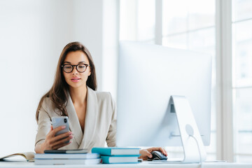 Elegant woman office worker uses mobile phone and computer at one time, sends messages and chats with clients online, monitors news from networks, sits at desktop with pile of notepads and books