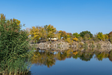 Autumn colors in sunny day on lake in city park. Landscape golden foliage and reflection of trees in calm water. Colorful plants on stone bank of pond and blue sky. Autumn reflections. Fall bright.