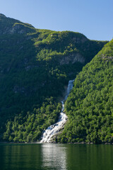 Waterfall in the Geiranger fjord surrounded by green trees and high mountains in Norway.