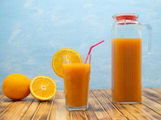 Glass and pitcher of orange juice on a wooden table on a blue background.
