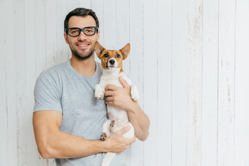Attractive cheerful Caucasian male in casual t shirt, spectacles, holds favourite pet, stands...