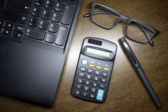 The glasses with the laptop , the calculator and the pen on the brown wooden working table.