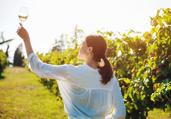 Young happy girl holding glass of white wine against sky and vineyard at sunset in sunlight