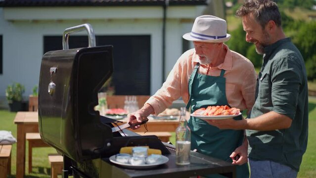 Senior father with adult son grilling outside on backyard in summer family during garden party