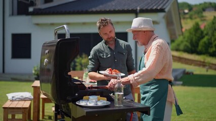 Senior father with adult son grilling outside on backyard in summer family during garden party