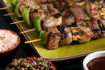 Macro close up of baked meat skewers on a green dish with sage leaves, salt and mixed pepper.